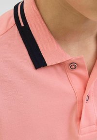 Šviesiai rožinė polo marškinėliai su tamsiai mėlynu dryžuotu apykakle, tekstūruotas audinys, dvi mygtukų eilės ir mažas logotipo detalė ant užsegimo.