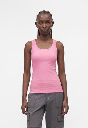 Femme aux cheveux tressés portant un débardeur rose et un pantalon cargo gris, debout devant un fond blanc uni.