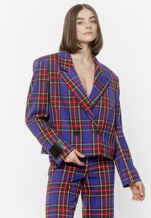 Kobieta w dużej niebiesko-czerwono-zielonej kratastej marynarce i dopasowanych spodniach, pozująca z ręką na biodrze na białym tle.