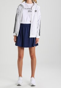 Vit rutig zip-up jacka, svart logotyp t-shirt, marinblå plisserad kjol, vita sportskor. Har djärva mönster och sportiga designelement.