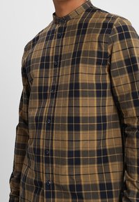 Button-up shirt in brown and navy plaid pattern, made of soft fabric, featuring a band collar and button closure along the front.