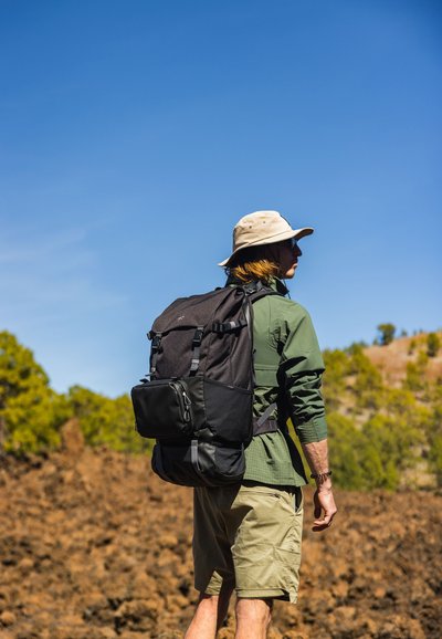 Mochila negra con correas ajustables y un bolsillo frontal, llevada por una persona con una camisa verde y pantalones cortos beige, con un cielo azul de fondo.
