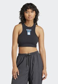 Svart ribbestrikket kort topp med en blå Adidas-etikett på brystet, i kombinasjon med løstsittende grå bukser. Enkelt design, uten ermer.