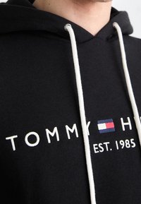 Svart hoodie med vita dragband, med broderad "Tommy Hilfiger" text, logotyp och "EST. 1985" på bröstet.