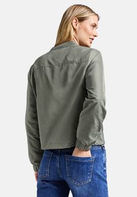 Donna con capelli biondi che indossa una giacca bomber verde oliva e jeans blu, girata di spalle con una mano nella tasca posteriore.