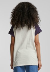 Cremefarvet og mørklilla t-shirt med korte ærmer og afslappet pasform, der har en rund halsudskæring og blød bomuldsstruktur, vist fra ryggen.