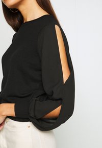 Suéter negro con un cuerpo de punto sólido y mangas abullonadas transparentes con detalles recortados. Los puños tienen un diseño recogido.