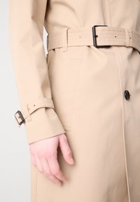 Mantou bej cu un design elegant, având o cataramă neagră la curea, închidere cu nasturi și un detaliu distinct la manșete.