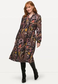 Langes schwarzes Kleid mit bunten Blumen- und Wirbelmustern, knöpfbare Vorderseite, lange Ärmel und taillierter Schnitt. Getragen mit kniehohen schwarzen Stiefeln.