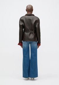 Καφέ δερμάτινο μπουφάν με ζώνη στη μέση και γιακά από γούνινη επένδυση, συνδυασμένο με φαρδιά τζιν παντελόνια σε μπλε απόχρωση. Η πίσω όψη δείχνει τις λεπτομέρειες και την εφαρμογή.