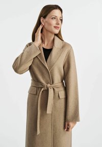 Beżowy długi płaszcz z wełny z paskiem w talii, kołnierzem typu szal i kieszeniami naszytymi. Gładka tekstura i prosty krój.