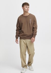 Sudadera marrón con un acabado texturizado, cuello redondo y texto blanco. Combinada con pantalones beiges y zapatillas blancas.