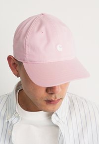 Boné de basebol de algodão rosa com aba curva, apresentando um logótipo branco na frente. Usado com uma camisa branca e uma camisa de botões às riscas.