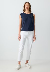 Top blu navy a maniche corte con dettaglio torcigliato sulla spalla sinistra, abbinato a pantaloni bianchi a gamba dritta e sneakers bianche.