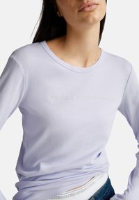 Frau trägt ein helllilanes Langarmshirt mit dem Schriftzug "United Colors of Benetton" auf der Brust, leicht in eine Blue Jeans gesteckt.