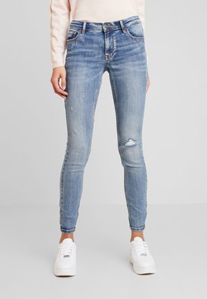 Femme portant un jean skinny bleu clair avec de petites déchirures et des baskets blanches, debout devant un fond gris clair uni.
