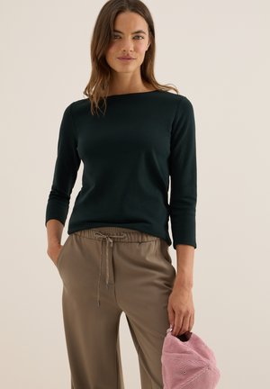 Haut à manches longues vert foncé avec un décolleté bateau, assorti à un pantalon beige avec cordon de serrage. Tenant une casquette en velours côtelé rose clair. Texture de tissu douce.