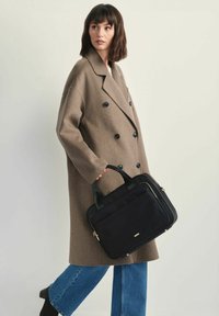 Kobieta ubrana w brązowy dwurzędowy płaszcz i niebieskie dżinsy, trzymająca czarną torebkę z złotymi detalami na jednolitym jasnym tle.