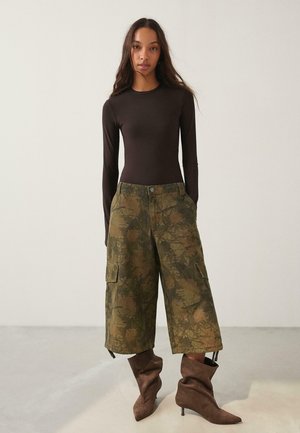 Frau trägt ein eng anliegendes langärmeliges schwarzes Oberteil, lockere Cargo-Hosen im Camouflage-Muster und braune lockere Stiefel mit Absatz, steht vor einem einfarbigen Hintergrund.