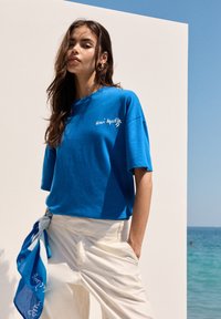 Blå bomulds T-shirt med afslappet pasform, med hvid tekst der står "Henri Matisse." Kombination med cremegule brede bukser og et blåt tørklæde.