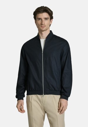 Young man with dark hair wears a navy zip-up jacket over a white shirt and beige pants, standing against a plain light background.
