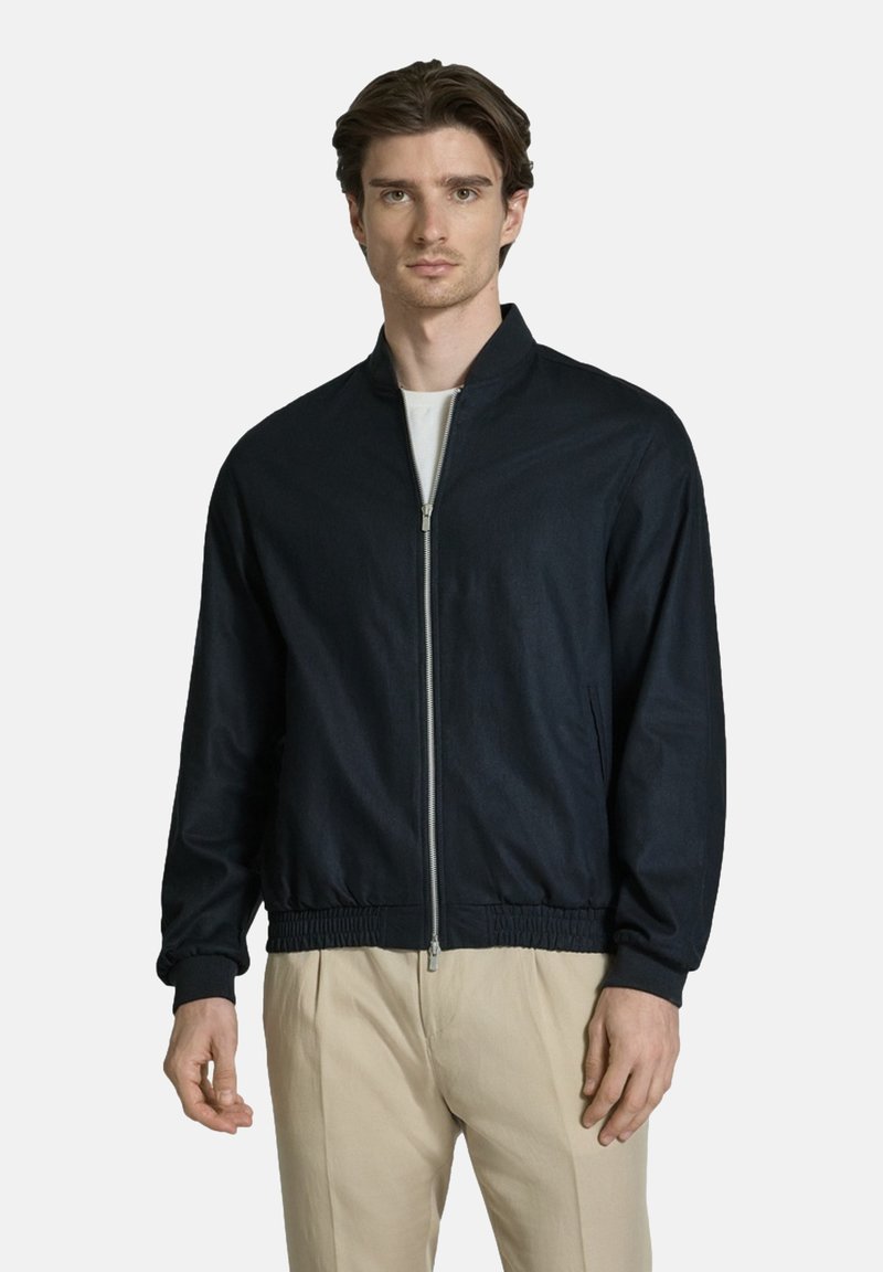 Young man with dark hair wears a navy zip-up jacket over a white shirt and beige pants, standing against a plain light background.