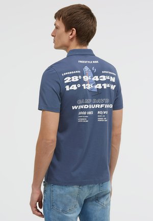 Man wearing blue short-sleeve polo with white windsurfing graphics and coordinates printed on the back, paired with light blue jeans.