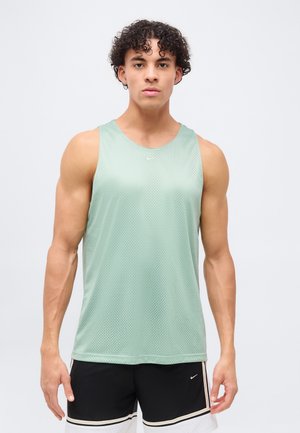 Junger Mann mit lockigem Haar, der ein hellgrünes ärmelloses Sportshirt und schwarz-weiße Shorts trägt, steht vor einem schlichten Hintergrund.