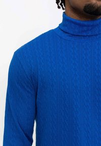 Pull à col roulé en tricot bleu avec un motif texturé en forme de diamant. Manches longues, matière douce et détail de col haut.