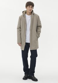 Vyras stovi vilkėdamas šviesiai rudos spalvos su gobtuvu striukę, baltą marškinėlį, tamsias džinsus ir juodus batus prieš paprastą baltą foną.