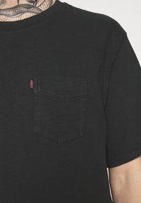 Czarny t-shirt z krótkim rękawem z teksturowanej tkaniny, z kieszonką na lewym piersiu i małą czerwoną metką na kieszeni.