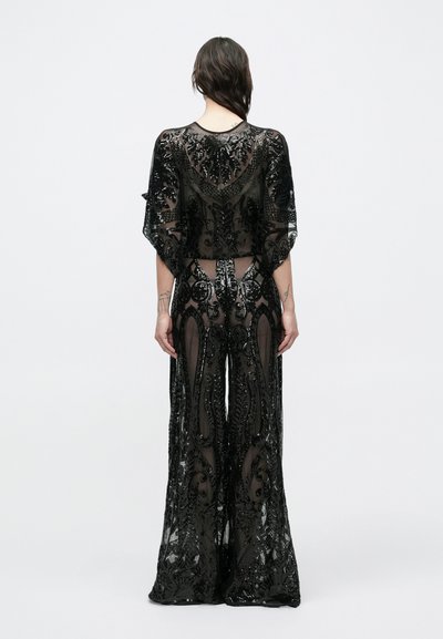 Nő áll háttal, áttetsző fekete díszített bő szárú nadrágot és hozzá illő áttetsző fekete, rövid ujjú hímzett felsőt visel.