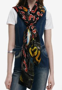Femme portant un t-shirt blanc et un gilet en jean avec une grande écharpe colorée à motifs nouée autour du cou, et un jean bleu.