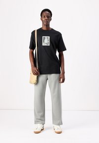 Sort t-shirt med et centralt grafisk motiv, grå brede bukser og hvide sneakers. En beige crossbody taske hænger over skulderen.