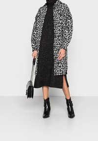 Cappotto a stampa leopardata in grigio e nero, abbinato a un vestito in maglia nera scintillante con spacchi laterali. Stivaletti neri e una piccola borsa.