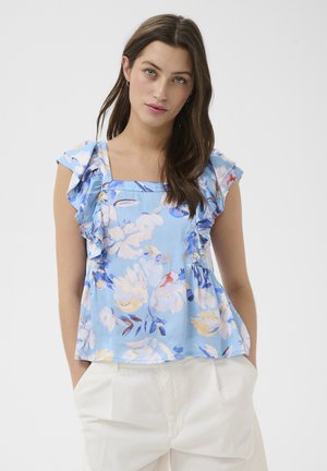 Femme portant un chemisier sans manches bleu à fleurs avec des volants sur les épaules et un pantalon blanc, debout devant un fond uni.