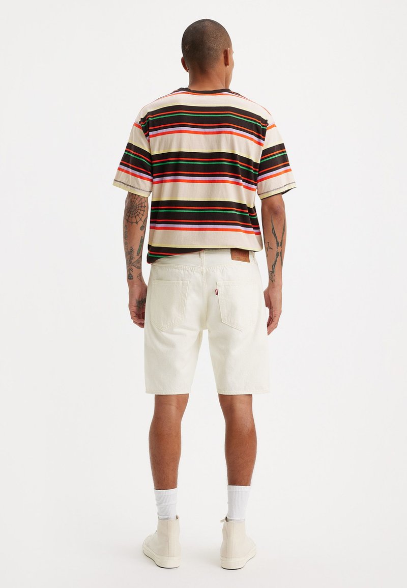 Kurzärmliges gestreiftes Hemd in Creme, Rot, Grün und Orange, kombiniert mit weißen Denim-Shorts. Sichtbares Markenlabel auf den Shorts.