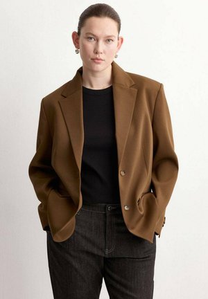 Mujer con blazer marrón, camisa negra y vaqueros oscuros, con las manos en los bolsillos, de pie frente a un fondo claro y sencillo.