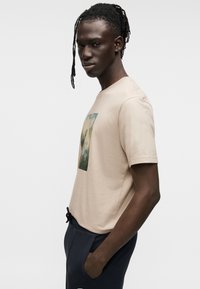 Μπεζ ανοιχτό βαμβακερό t-shirt με ορθογώνιο γραφικό σχέδιο σε πράσινο και μπλε. Διαθέτει στρογγυλή λαιμόκοψη και κοντά μανίκια. Κανονική εφαρμογή.