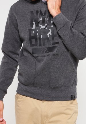 Homme portant un sweat à capuche gris foncé avec le texte "NYC BIKE PRL" et un pantalon beige, main près du visage, corps partiellement visible.