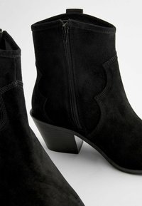 Black suede ankle boots with a low heel, featuring a side zipper and curved stitching details. Smooth texture, structured design.