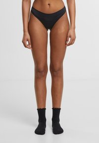 Fekete bikini alsó sima textúrával és magas vágású dizájnnal, fekete bordázott zoknikkal kombinálva, fodros tetejű szélekkel.