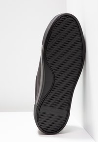 Semelle d'une basket noire avec motif de grip texturé et marquage subtil, positionnée à la verticale contre un coin blanc.