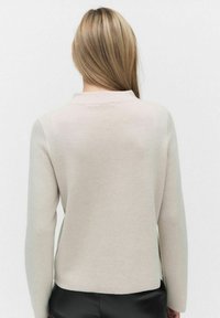 Beige neulepaita, jossa on teksturoitu neulos, pitkät hihat, pyöreä kaula-aukko ja hieman lyhennetty helma. Saumayksityiskohdat selässä lisäävät hienovaraista mielenkiintoa.
