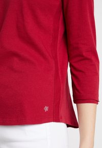 La camicia rossa di cotone a maniche lunghe presenta cuciture laterali e un orlo che curva verso l'alto ai lati. Un piccolo logo è posizionato vicino in basso a sinistra.