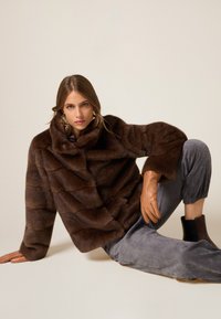 Braune Kunstpelzjacke mit strukturiertem wellenförmigem Muster, übergroßem Kragen und Knopfverschluss. Kombiniert mit grauen Cordhosen und Lederhandschuhen.
