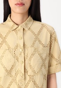 Camicia gialla con ritagli a forma di diamante, chiusura a bottoni e due tasche sul petto. Il tessuto ha una finitura testurizzata con un branding discreto.