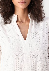 Vit broderad blus med intrikata utskärningsmönster, en V-ringning och en texturerad yta. Mjuk tyg med en delikat design.