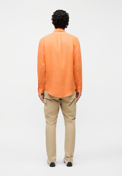 Άνδρας με σγουρά μαλλιά, ντυμένος με πορτοκαλί πουκάμισο μακρυμάνικο και μπεζ παντελόνι φόρμας cargo, στέκεται με την πλάτη στραμμένη και τα χέρια χαλαρά στο πλάι.
