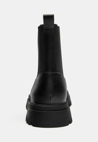 Čierna kožená členková čižma s hrubou podrážkou a elastickou bočnou vložkou, fotografovaná zozadu na jednoduchom svetlom pozadí.